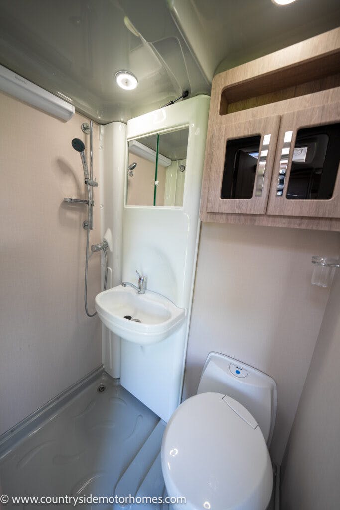 A compact motorhome bathroom in the 2021 Auto-Sleepers Broadway EL featuring a shower, a small sink with a faucet, and a mirror above. There's also a toilet and a wooden cabinet with mirrored doors on the wall. The floor is designed for wet use. Website text is visible at the bottom.