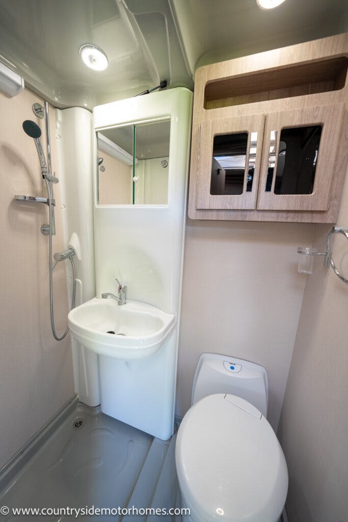 A compact bathroom in the 2021 Auto-Sleepers Broadway EL features a small sink with a mirror above, a showerhead, a wall-mounted wooden cabinet, and a toilet. The walls and fixtures are in neutral tones, and there is also a towel ring on the right wall.