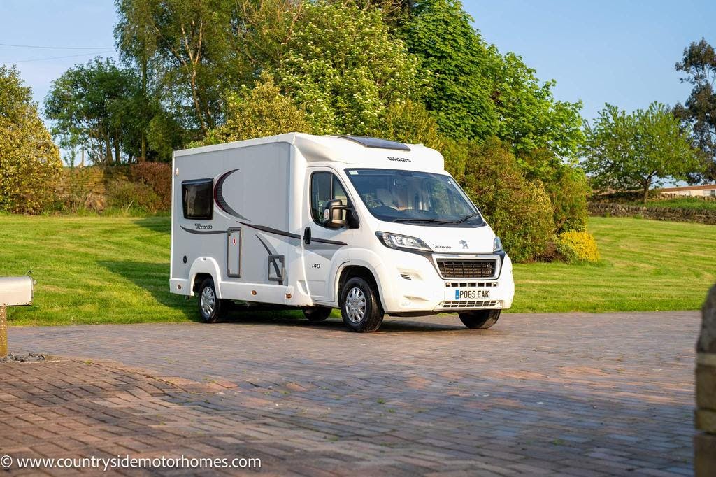 A 2015 Elddis Accordo 140 campervan with "Elddis" branding is parked on a paved driveway surrounded by lush green grass, trees, and bushes. The countryside setting is peaceful and idyllic. The campervan has a UK license plate that reads "PX08 EAK.